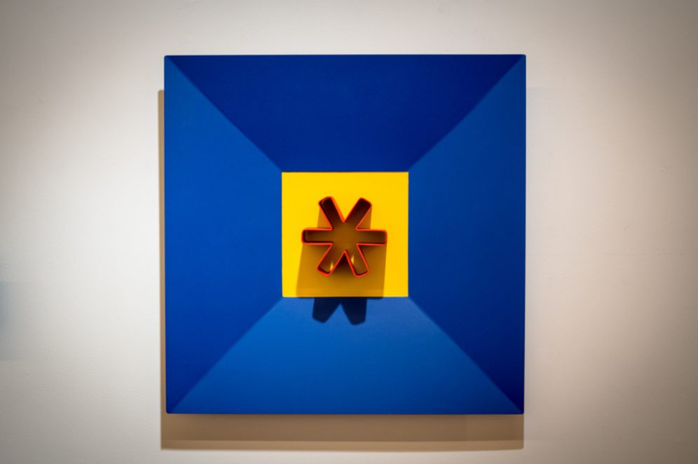 Escultura de asterisco montada sobre un panel azul y amarillo, en pared
